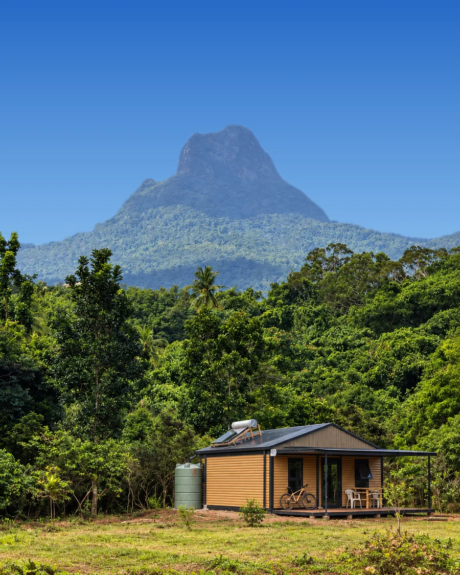 An off-grid cabin with solar panels in a rainforest clearing, below a twin-peaked mountain